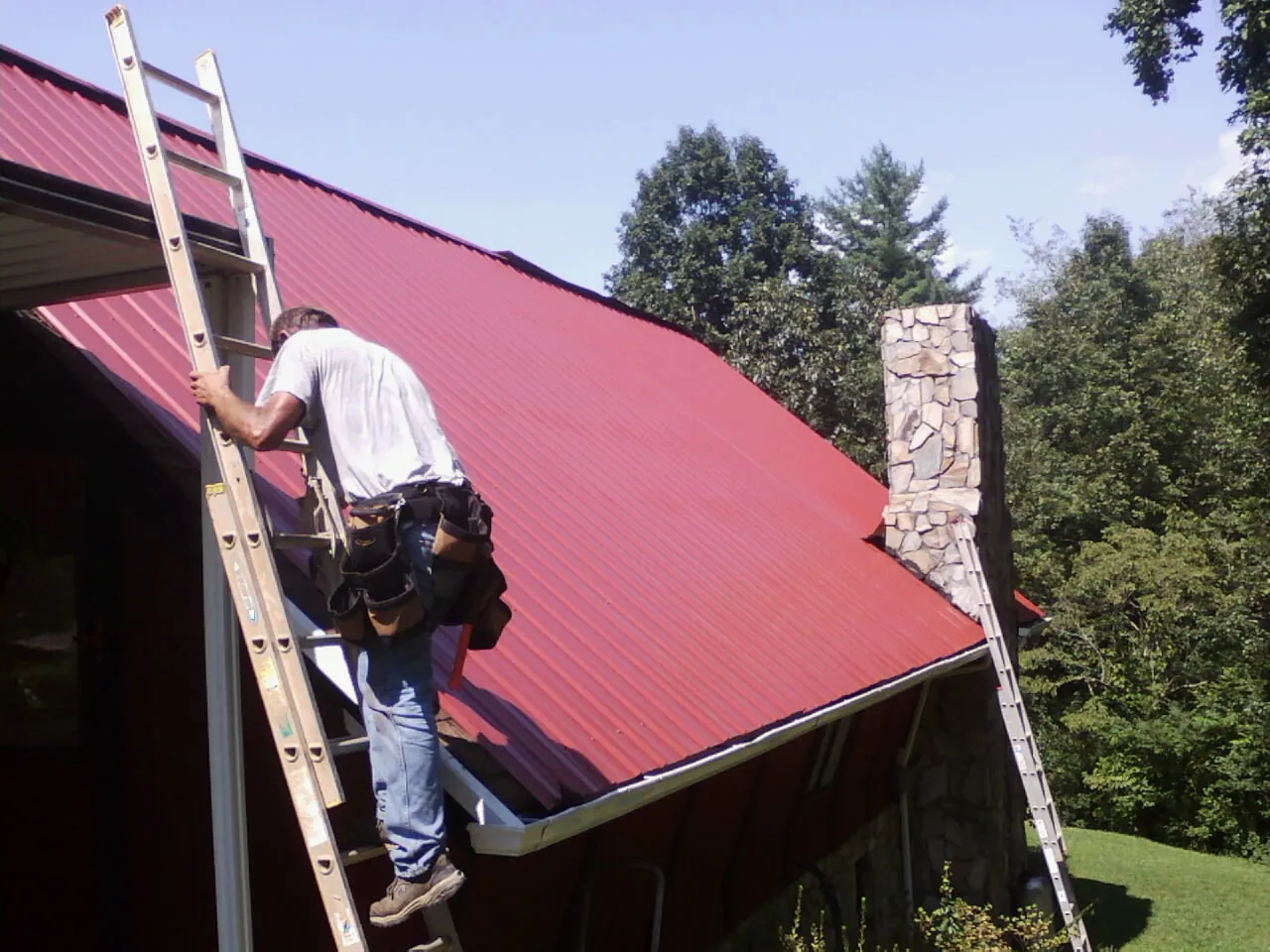 Roofer on a ladder beside a red metal roof and stone chimney — Soffit & Fascia Repair by Roofing Brothers in Rowland Heights